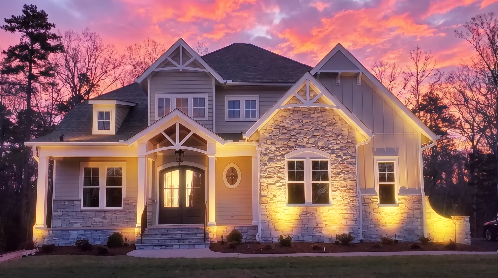 Two story stone and siding home at dusk with warm white landscape uplights on the facade, installed by Green Light